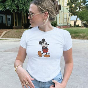 May include: White t-shirt with a Mickey Mouse graphic. The cartoon character is shown in red shorts, brown shoes, and black gloves. A classic design for Disney fans, perfect for casual wear.
