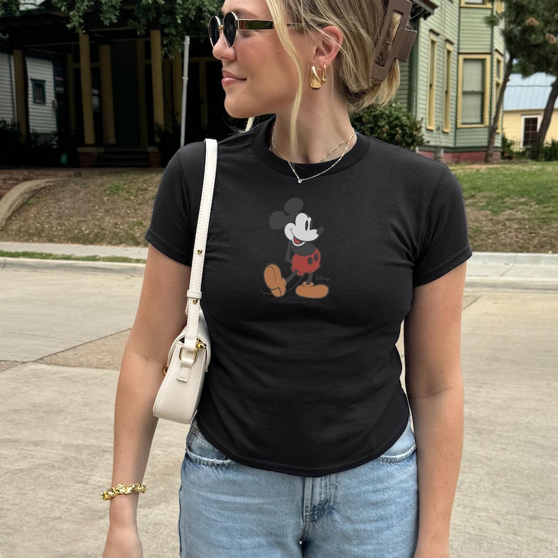 May include: Black crew neck t-shirt with a Mickey Mouse graphic. The cartoon character is shown in red shorts, yellow shoes, and white gloves. The shirt is worn with light blue denim jeans.