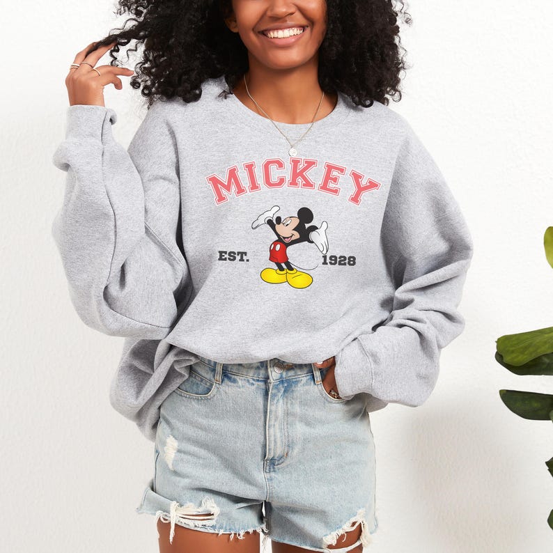 May include: A gray sweatshirt featuring the word "MICKEY" in pink lettering above a graphic of Mickey Mouse. Below the graphic are the words "EST. 1928". The sweatshirt has long sleeves and a crew neck.