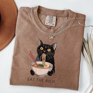 Może przedstawiać: Brązowa koszulka z czarnym kotem jedzącym ramen. Kot patrzy na widza, a pod nim widnieje napis "EAT THE RICH".