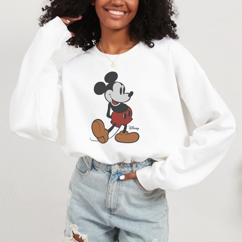 May include: White sweatshirt featuring a classic Mickey Mouse graphic. The design showcases Mickey in his iconic red shorts, black shorts, and brown shoes. The Disney logo is printed below the character. The sweatshirt is a casual style.