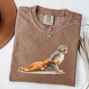 May include: A brown Comfort Colors t-shirt featuring a graphic of a gray monkey carrying a brown teddy bear. The shirt has a crew neck and short sleeves. A silver necklace with a small pendant is draped across the shirt.