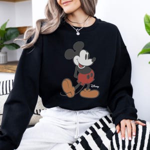 Pu&ograve; includere: Felpa nera con grafica di Topolino. Il personaggio dei cartoni animati &egrave; raffigurato nella sua posa classica, con pantaloncini rossi, scarpe marroni e guanti bianchi. Il logo Disney &egrave; stampato in basso a destra.