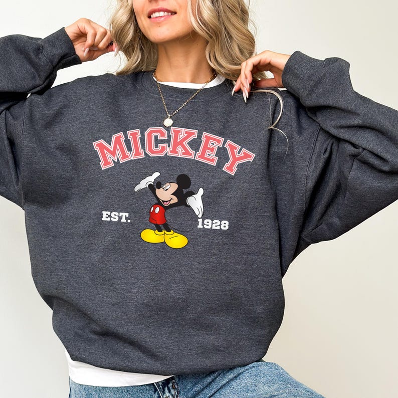 May include: Dark gray sweatshirt featuring the word "Mickey" in pink lettering above a cartoon image of Mickey Mouse. The sweatshirt also includes the text "EST. 1928". The design is centered on the front of the garment.