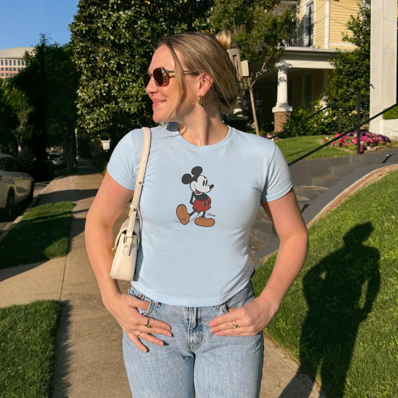 May include: A pale blue t-shirt with a Mickey Mouse design. The cartoon character is shown in his classic pose, wearing red shorts and brown shoes. The shirt is worn with light blue denim jeans.