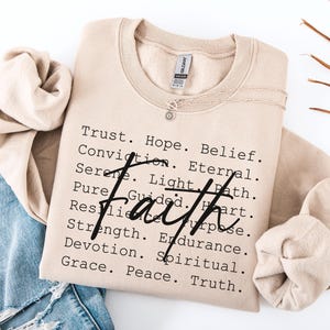 May include: A beige crewneck sweatshirt with the word "Faith" in a bold, black script. The sweatshirt features a list of words like "Trust," "Hope," and "Belief." A delicate silver necklace with a small pendant rests on the fabric. The sweatshirt is folded with a pair of blue jeans.
