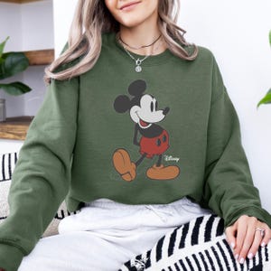 Pu&ograve; includere: Una felpa verde oliva con una grafica di Topolino. Il personaggio dei cartoni animati &egrave; raffigurato nella sua posa classica, con pantaloncini rossi e scarpe marroni. Il logo Disney &egrave; visibile in basso a destra.