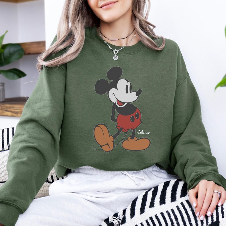 Pu&ograve; includere: Una felpa verde oliva con una grafica di Topolino. Il personaggio dei cartoni animati &egrave; raffigurato nella sua posa classica, con pantaloncini rossi e scarpe marroni. Il logo Disney &egrave; visibile in basso a destra.
