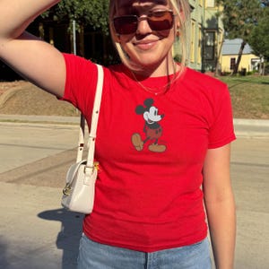 May include: A red t-shirt with a graphic of Mickey Mouse. The short-sleeved shirt has a crew neck. The Mickey Mouse graphic is in black, red, yellow, and tan. The shirt is worn with blue jeans. A small, white handbag with a gold clasp is also visible.