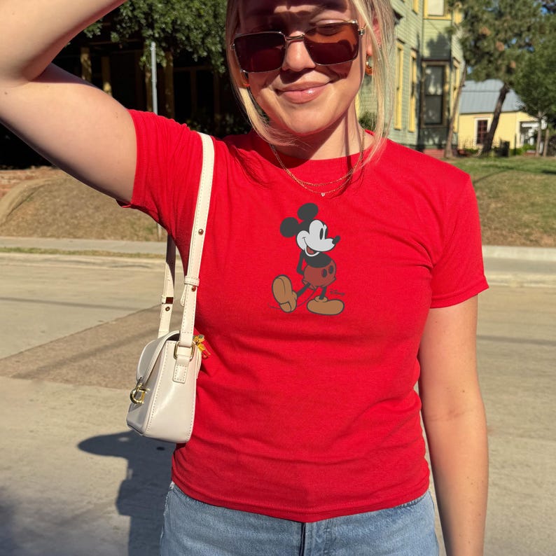 May include: A red t-shirt with a graphic of Mickey Mouse. The short-sleeved shirt has a crew neck. The Mickey Mouse graphic is in black, red, yellow, and tan. The shirt is worn with blue jeans. A small, white handbag with a gold clasp is also visible.