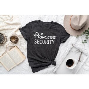 Puede incluir: Una camiseta gris oscuro con un gr&aacute;fico blanco que dice "Princess Security" en una fuente estilizada.