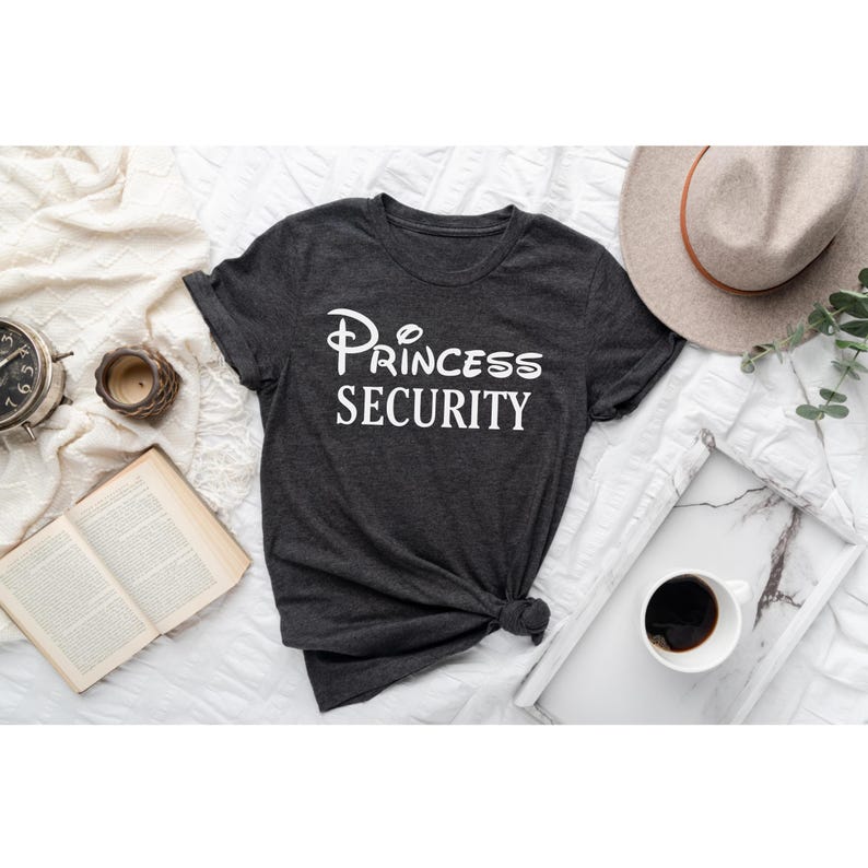 Puede incluir: Una camiseta gris oscuro con un gr&aacute;fico blanco que dice "Princess Security" en una fuente estilizada.