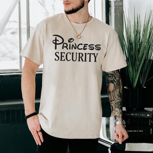 Puede incluir: Una camiseta beige con el texto "Princess Security" en letras negras. El texto est&aacute; estilizado para parecerse al logotipo de Disney.