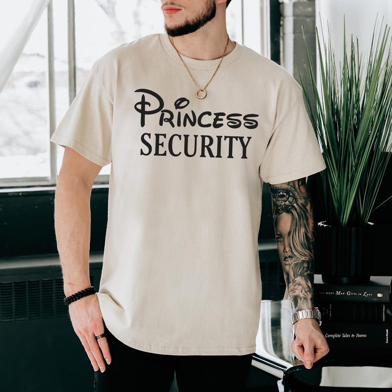Puede incluir: Una camiseta beige con el texto "Princess Security" en letras negras. El texto est&aacute; estilizado para parecerse al logotipo de Disney.