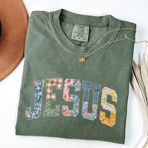 Puede incluir: Una camiseta verde oliva con la palabra "JESUS" aplicada en tela de retazos. El patchwork incluye patrones florales, a cuadros y lisos. Un collar plateado con un pequeño colgante descansa sobre la camiseta.