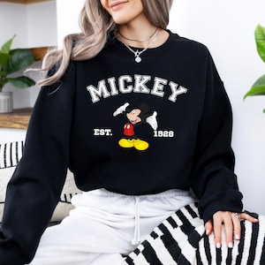 May include: Black sweatshirt featuring the word "MICKEY" in white block letters above a cartoon image of Mickey Mouse. The image includes the text "EST. 1928". The sweatshirt is a classic crew neck style.