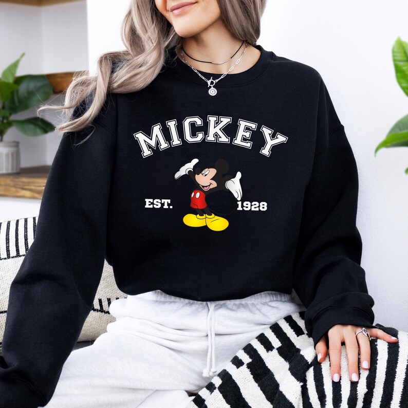May include: Black sweatshirt featuring the word "MICKEY" in white block letters above a cartoon image of Mickey Mouse. The image includes the text "EST. 1928". The sweatshirt is a classic crew neck style.