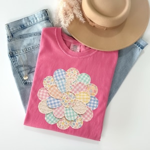 以下が含まれることがあります： 様々な柄の生地で作られた、大きなカラフルな花の模様が特徴のピンク色のTシャツ。青いジーンズとベージュの帽子と組み合わせています。花の模様にはギンガムチェックと花柄が含まれています。