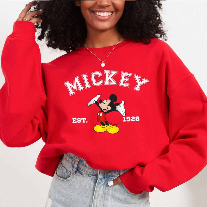 May include: A red sweatshirt featuring the word "MICKEY" in white block letters above a cartoon image of Mickey Mouse. Below the image are the words "EST. 1928". The sweatshirt is a solid red color.