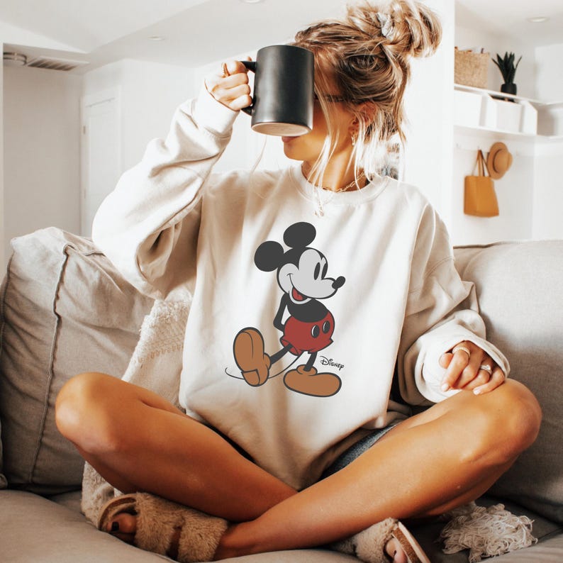 Pu&ograve; includere: Una felpa beige con una grafica di Topolino. Il personaggio dei cartoni animati &egrave; raffigurato nella sua posa iconica, con orecchie nere, viso bianco, pantaloncini rossi e scarpe marroni. Il logo Disney &egrave; stampato sotto.