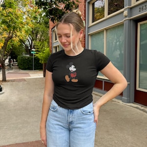 May include: Black short-sleeved t-shirt with a Mickey Mouse graphic. The cartoon character is in black, white, red, and tan. The shirt is worn with light blue denim jeans. The person is standing outside on a pavement.