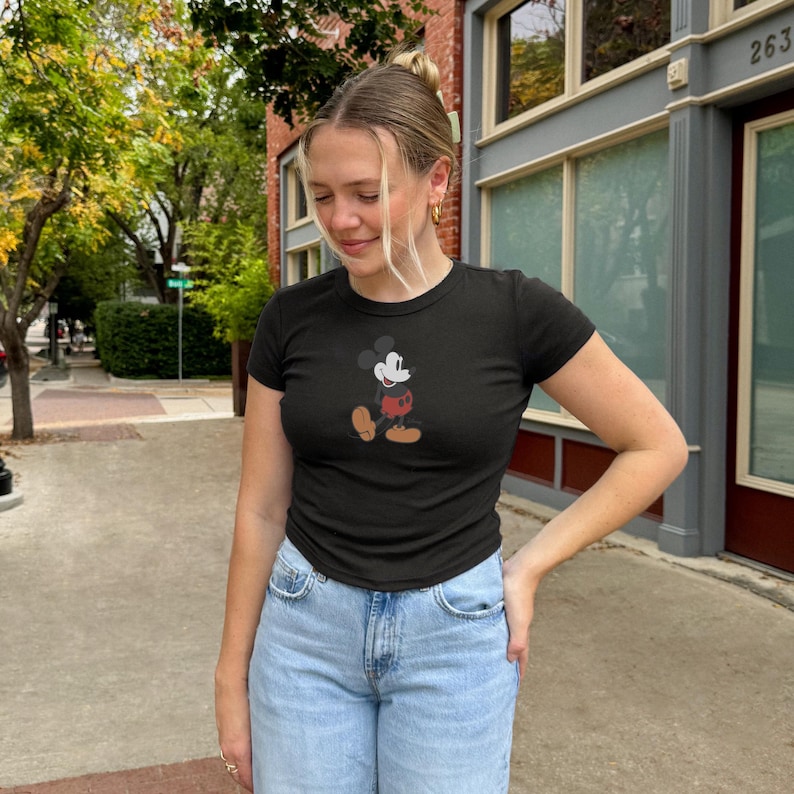 May include: Black short-sleeved t-shirt with a Mickey Mouse graphic. The cartoon character is in black, white, red, and tan. The shirt is worn with light blue denim jeans. The person is standing outside on a pavement.