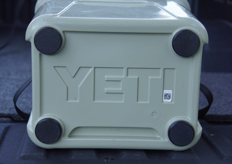 May include: A light green cooler with the brand name "YETI" embossed on the front. The cooler has four black rubber feet and a black strap. A small QR code sticker is visible.