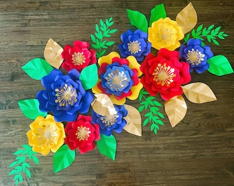 Könnte beinhalten: Eine bunte Papierblumen-Arrangement mit blauen, roten, gelben und goldenen Blumen. Die Blumen sind auf einem braunen Holzgrund angeordnet.