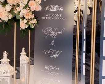 May include: A frosted glass sign with a gold frame announcing a wedding. The sign reads "Welcome to the Nikkah of Rabiyah & Bilal" and includes an Arabic inscription and the date "October 6, 2024".