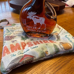 May include: A glass bottle of pure maple syrup with a moose design. The bottle is sitting on a white fabric bag with a map of New Hampshire and the text "The Granite State".
