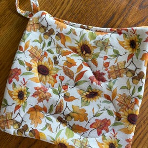 May include: A square quilted potholder featuring a white background with a repeating pattern of yellow sunflowers, orange and red autumn leaves, and brown acorns. The fabric item includes a small fabric loop at the top corner for hanging.