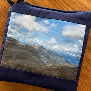 May include: A navy blue pot holder with a mountain landscape printed on the front. The landscape features a mountain range with a blue sky and white clouds.