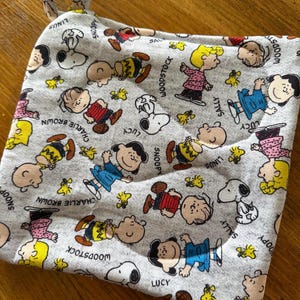May include: A square fabric pot holder with a gray heather background featuring a repeating pattern of Peanuts characters including Snoopy, Charlie Brown, Lucy, Linus, Sally, and Woodstock, with their names printed next to them and a fabric loop.