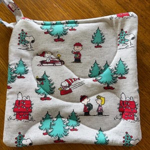 May include: A square quilted pot holder in a light gray fabric featuring a repeating Peanuts holiday print. The pattern includes Snoopy, Charlie Brown, and Woodstock among teal Christmas trees, red doghouses with lights, and sledding scenes.
