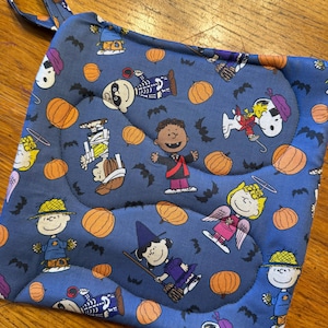 May include: A square quilted pot holder in blue fabric featuring a repeating Halloween pattern of Peanuts characters in costumes, including Snoopy, Charlie Brown, and Lucy, interspersed with orange pumpkins and small black bats across the surface.