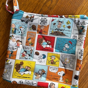 May include: A square fabric pot holder featuring a colorful Peanuts comic strip print with Snoopy, Charlie Brown, and other characters. The design includes various sports scenes, speech bubbles with text like "POW!", "AAUGH!", and "STRIKE ONE!", and a fabric loop for hanging.