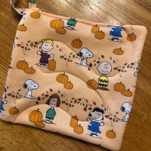 May include: Peach-colored, quilted pot holder with a loop for hanging. The fabric features a repeating pattern of orange pumpkins, falling leaves, and cartoon characters. The pot holder is square and has a stitched design.