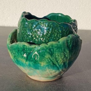 Puede incluir: Dos cuencos de cerámica verde con forma de col. Los cuencos están anidados, con el cuenco más pequeño encima del más grande.