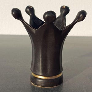 Puede incluir: Objeto decorativo en forma de corona, de color marrón oscuro. Tiene una banda dorada en la base y cinco puntas que se extienden hacia arriba con extremos redondeados. Hecho de un material liso, posiblemente metálico.