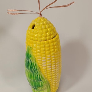 Op de afbeelding: Een keramische maïs-vormige container met een deksel. De container is geel met witte korrels en een groene schil. Het deksel heeft een klein gaatje bovenaan en een bruin lint is eromheen gebonden.