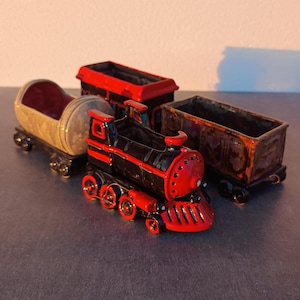 Può includere: Un set di treni giocattolo in ceramica, con una locomotiva nera e rossa, un vagone merci e un vagone di coda. Il treno ha uno stile vintage.