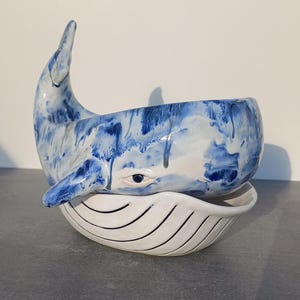 May include: A ceramic whale bowl with a blue and white glaze. The whale is depicted with a single eye and a flowing tail. The bowl is nested inside a smaller, white bowl with blue stripes.