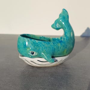May include: A turquoise ceramic whale planter with a white base. The whale has a detailed eye and a raised tail. The planter is designed to hold small plants or succulents, adding a whimsical touch to any space.