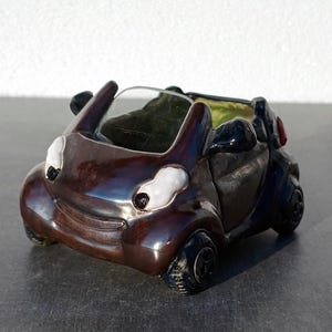 May include: A brown ceramic car planter with white eyes and a black windshield. The car has a black interior and black wheels.