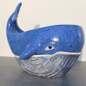 May include: A blue ceramic whale bowl with a white eye and a textured surface. The bowl is sitting on a smaller white bowl with a blue and white wave pattern.