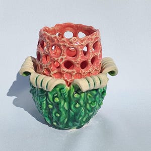 May include: A ceramic planter with a green base and a pink, open-work top. The planter is shaped like a flower with a ruffled edge.