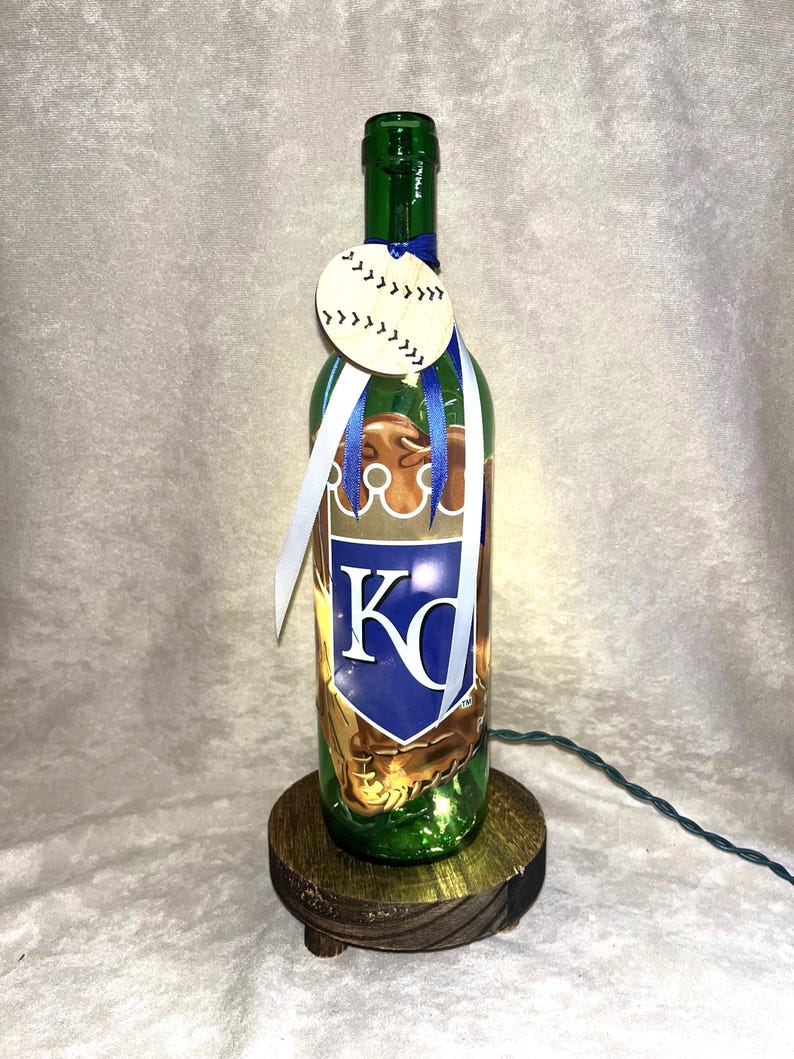 Puede incluir: Una botella de vidrio verde reutilizada como l&aacute;mpara, con una etiqueta de madera con tem&aacute;tica de b&eacute;isbol y cintas. La botella tiene un logotipo del equipo con las letras "KC" en azul y blanco. Se encuentra sobre una peque&ntilde;a base de madera redonda.