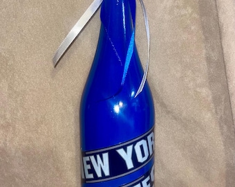 Puede incluir: Una botella azul cobalto con "NEW YORK YANKEES" impreso, coronada con una pelota de béisbol. Cintas blancas y azules atadas alrededor del cuello. Un artículo decorativo para los fanáticos del deporte.