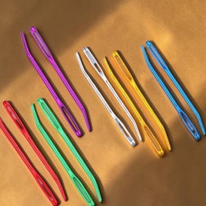 Puede incluir: Una colección de agujas de lana coloridas en varios tonos, incluyendo rojo, verde, morado, plateado, amarillo y azul. Las agujas son de metal y tienen ojos grandes para facilitar el enhebrado. Ideales para proyectos de manualidades y costura.