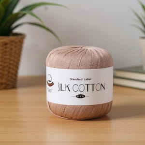 May include: A ball of silk cotton yarn in a light brown color. The yarn has a label that reads "Standard Label SILK COTTON". The yarn is sitting on a wooden surface.